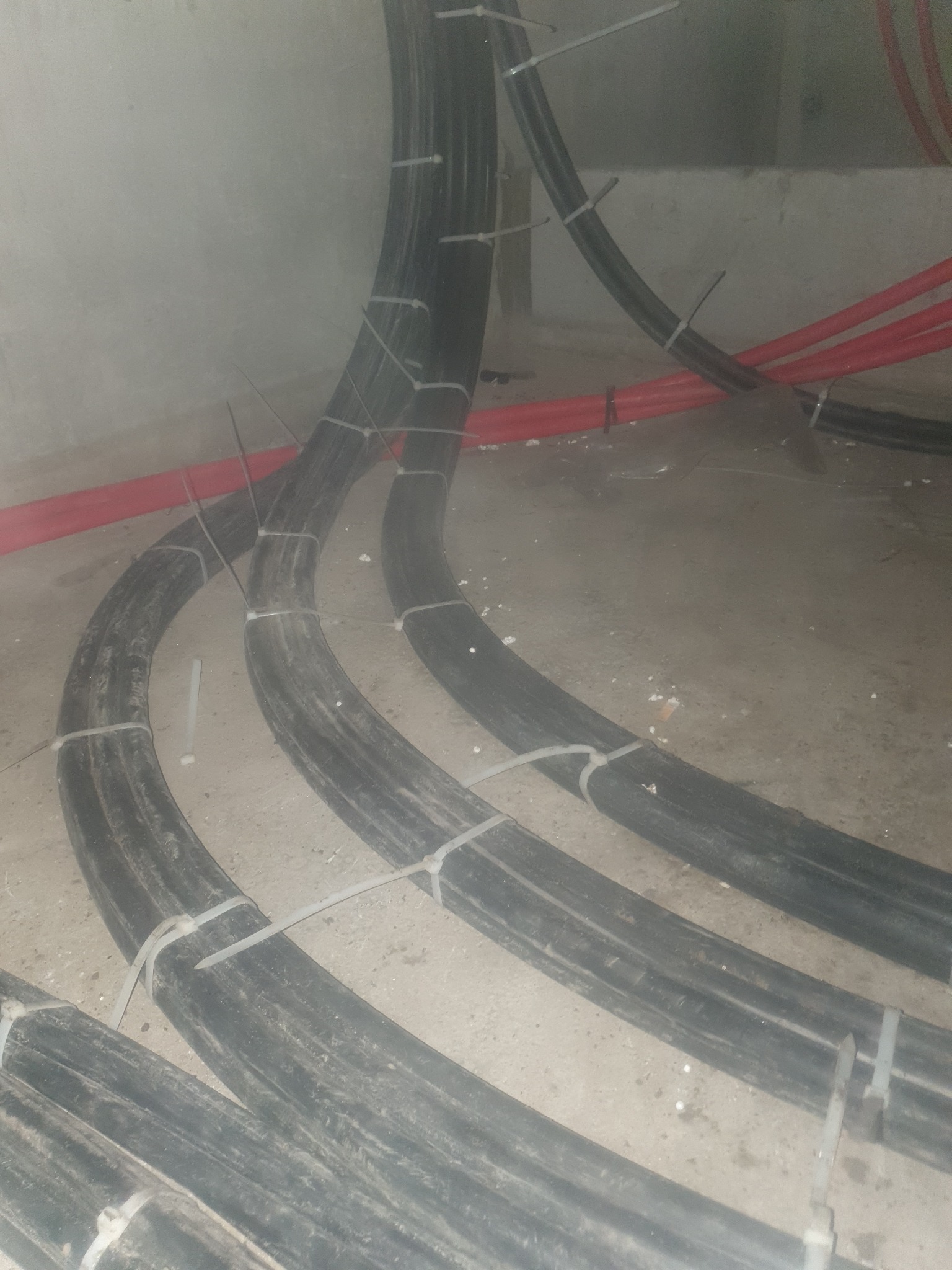 Grube kable elektryczne czarne i czerwone, ułożone łukami i spięte białymi opaskami zaciskowymi na betonowej posadzce w szarym pomieszczeniu.