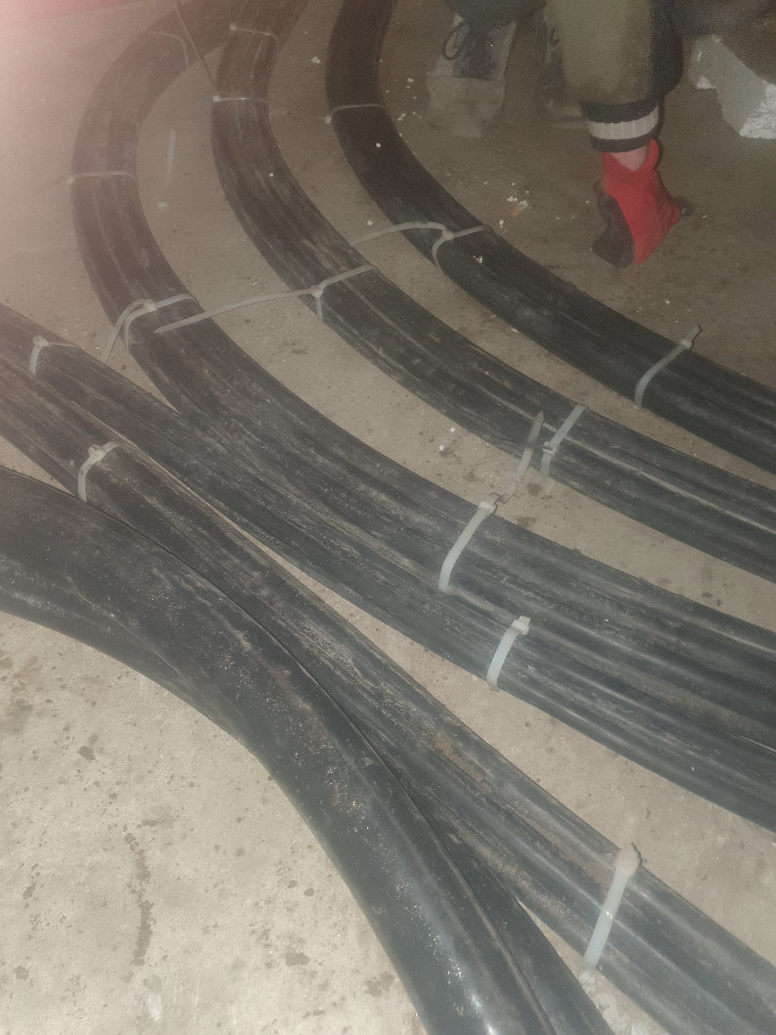 Układ kilku grubych, czarnych kabli elektrycznych, spiętych białymi opaskami zaciskowymi, na szarym, betonowym podłożu; fragment nogi w czerwonej skarpecie widoczny w tle.