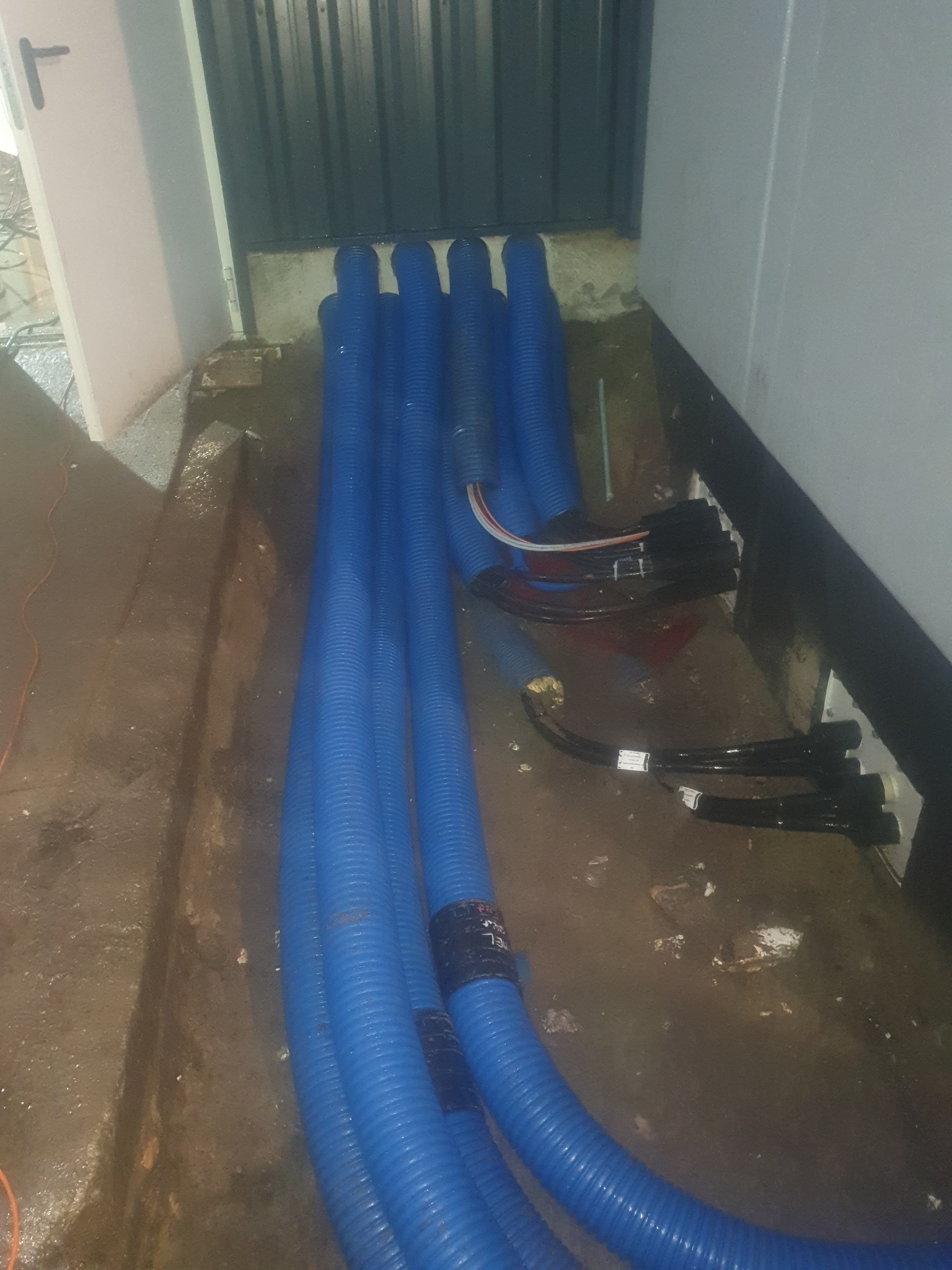 Rury karbowane w kolorze niebieskim i przewody elektryczne leżące na podłodze w pobliżu ściany i metalowych drzwi garażowych.