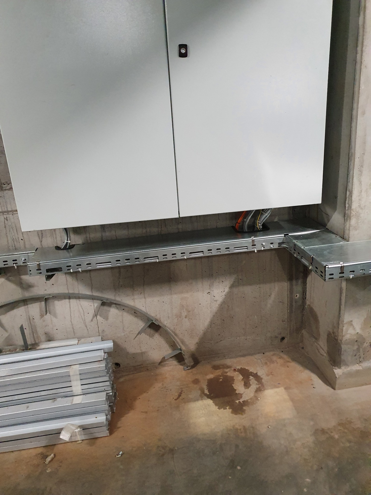 Szara szafa elektryczna zamontowana na betonowej ścianie, z widocznymi kablami w metalowej korycie kablowym i stosem nieużywanych koryt na podłodze. Widoczna plama na betonowej podłodze.