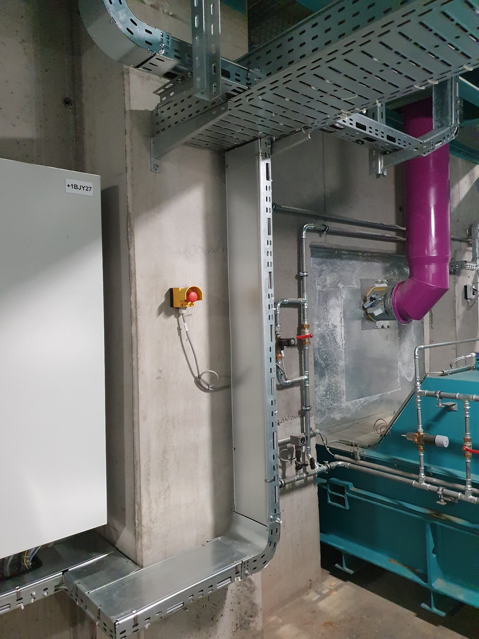 Szare koryta kablowe, rury i skrzynka elektryczna na tle betonowej ściany z przyciskiem awaryjnym w Reskowie.