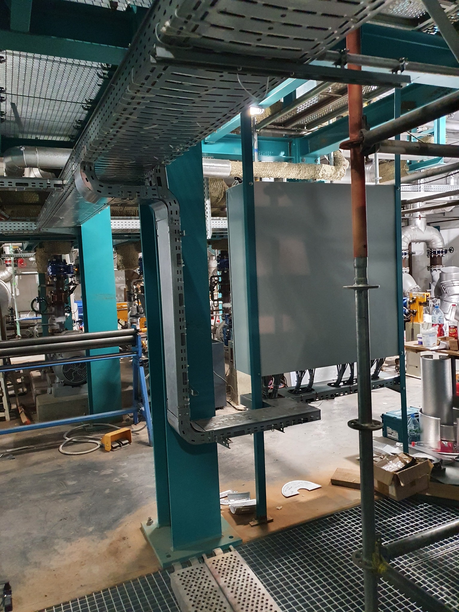 Widok na metalowe koryto kablowe przymocowane do zielonej konstrukcji wsporczej w pomieszczeniu przemysłowym, z widocznymi rurami i elementami instalacji w tle. Na podłodze widoczne kraty pomostowe...