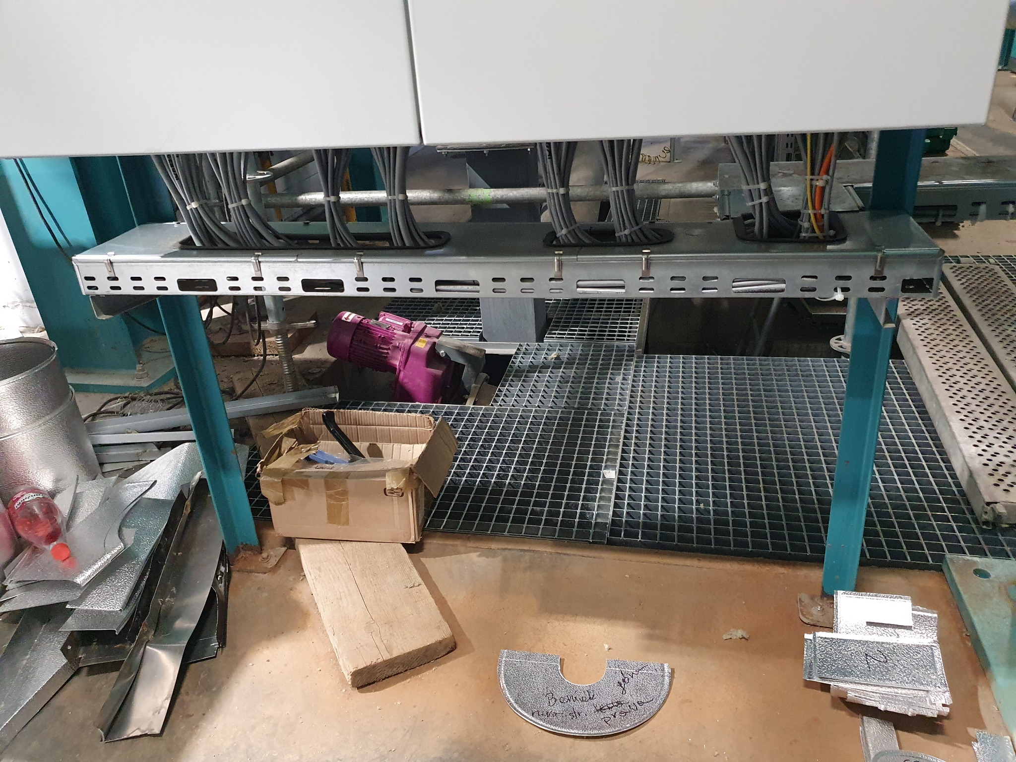 Szare przewody elektryczne w metalowych korytach podwieszonych pod białą obudową, widoczne elementy konstrukcji stalowej, na podłodze resztki materiałów i narzędzi.