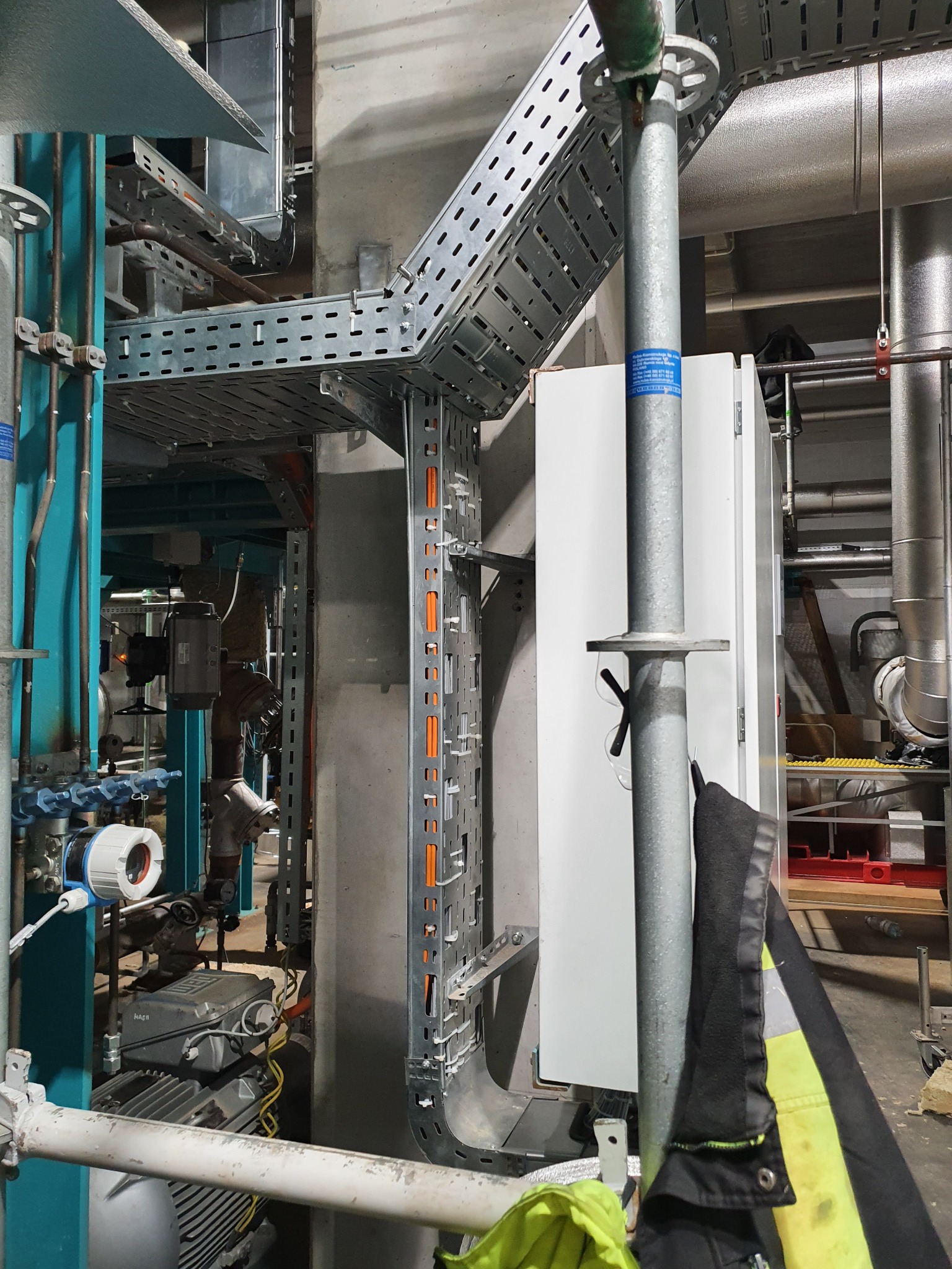 Metalowe korytka kablowe prowadzone wzdłuż betonowej ściany w hali przemysłowej, widoczne rury, instalacje oraz szafa sterownicza w tle, na pierwszym planie fragment rusztowania i odzież robocza.