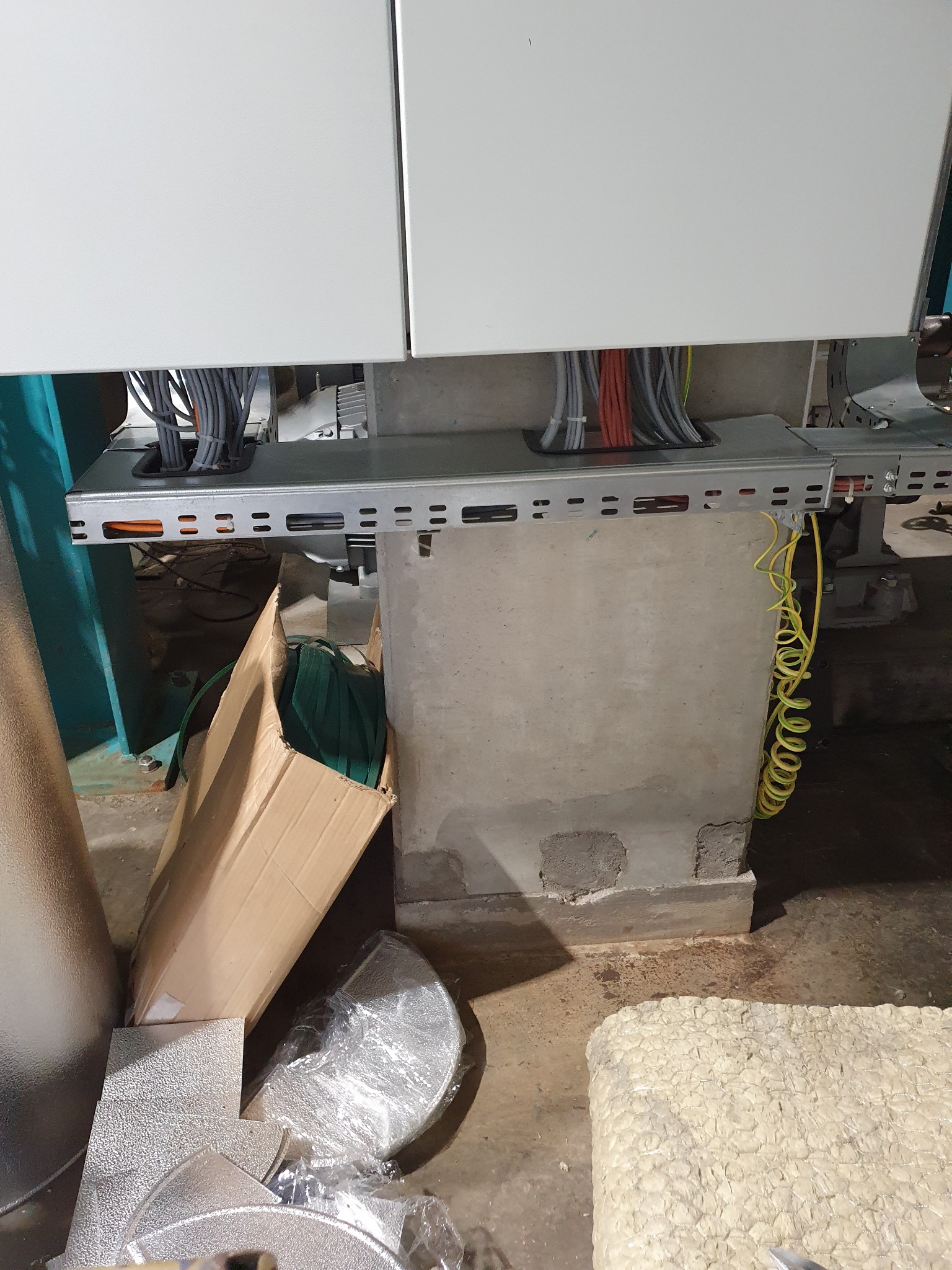 Szara metalowa korytka kablowa z ułożonymi przewodami elektrycznymi wychodzącymi z białej szafy, z żółto-zielonym przewodem spiralnym zwisającym wzdłuż betonowego filara. Na podłodze widoczne...