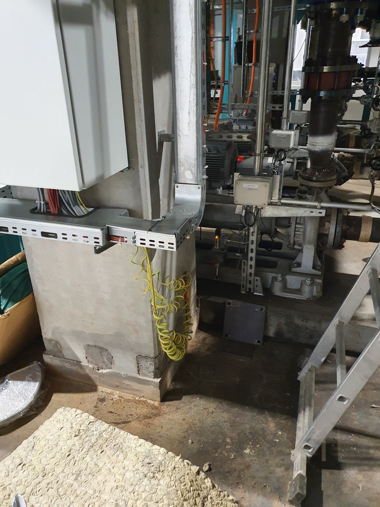Szara betonowa kolumna z zamontowanymi metalowymi korytkami kablowymi, w których ułożone są przewody elektryczne o różnych kolorach, w tle przemysłowe rury i drabina.