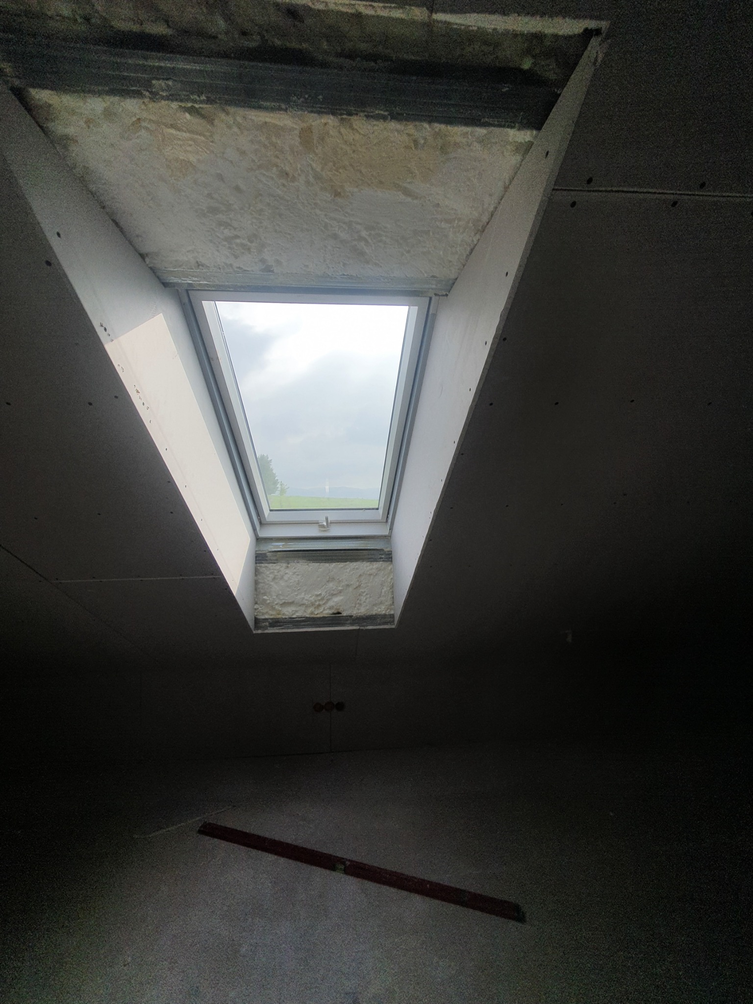 Wnętrze poddasza w trakcie prac wykończeniowych; widoczne okno dachowe z widokiem na zewnątrz, płyty gipsowo-kartonowe na ścianach i suficie, izolacja wokół okna, poziomica na podłodze w słabym...