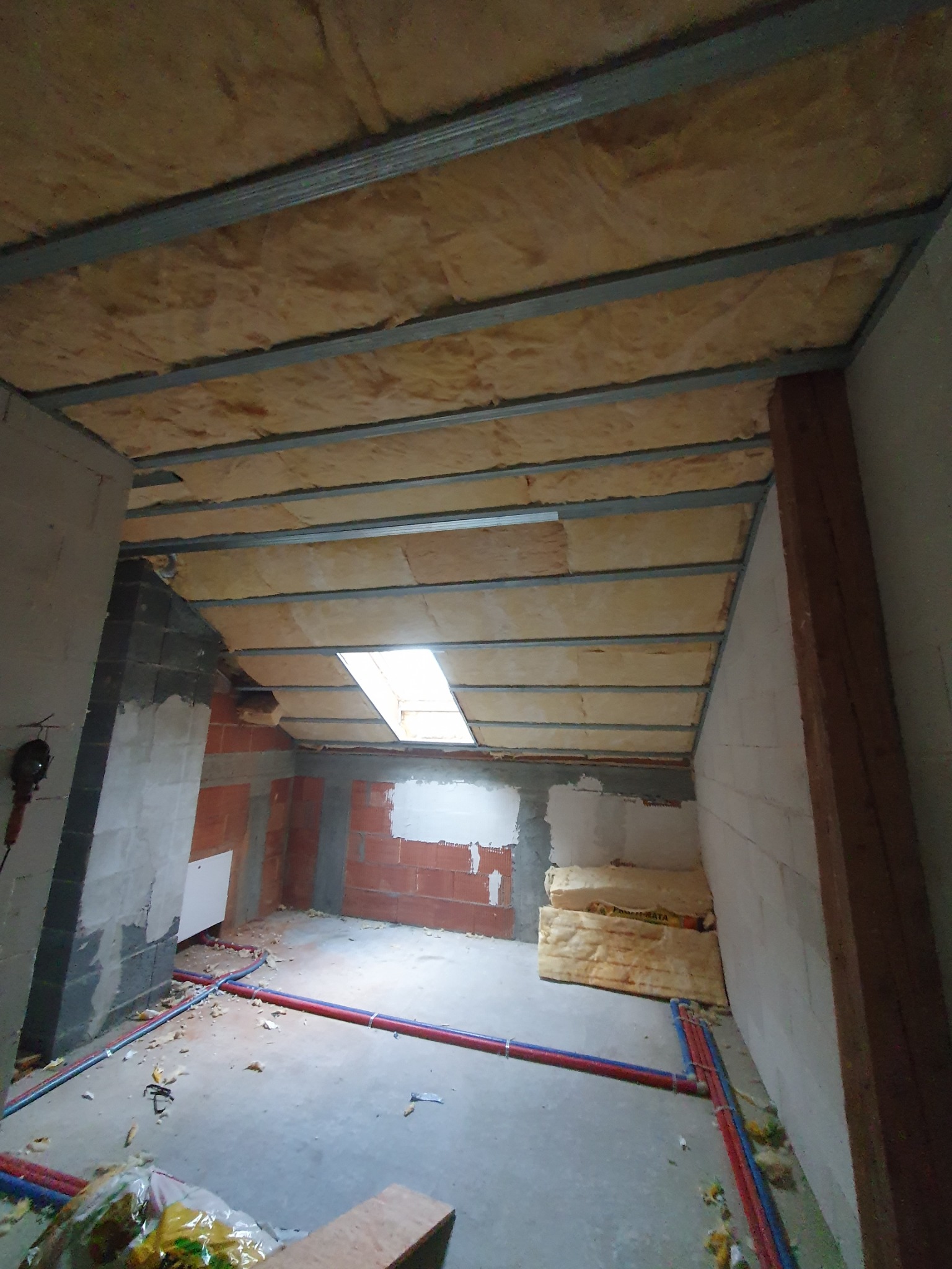 Poddasze w trakcie prac wykończeniowych: widoczne ocieplenie wełną mineralną między metalowymi profilami, fragment ściany z cegły, rury instalacyjne na podłodze, okno dachowe, elementy...