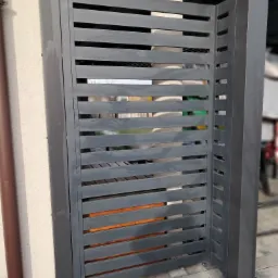 Nowoczesna, szara brama wjazdowa z poziomych profili, zamontowana na posesji z kostką brukową, widoczny fragment rynny w kolorze brązowym.