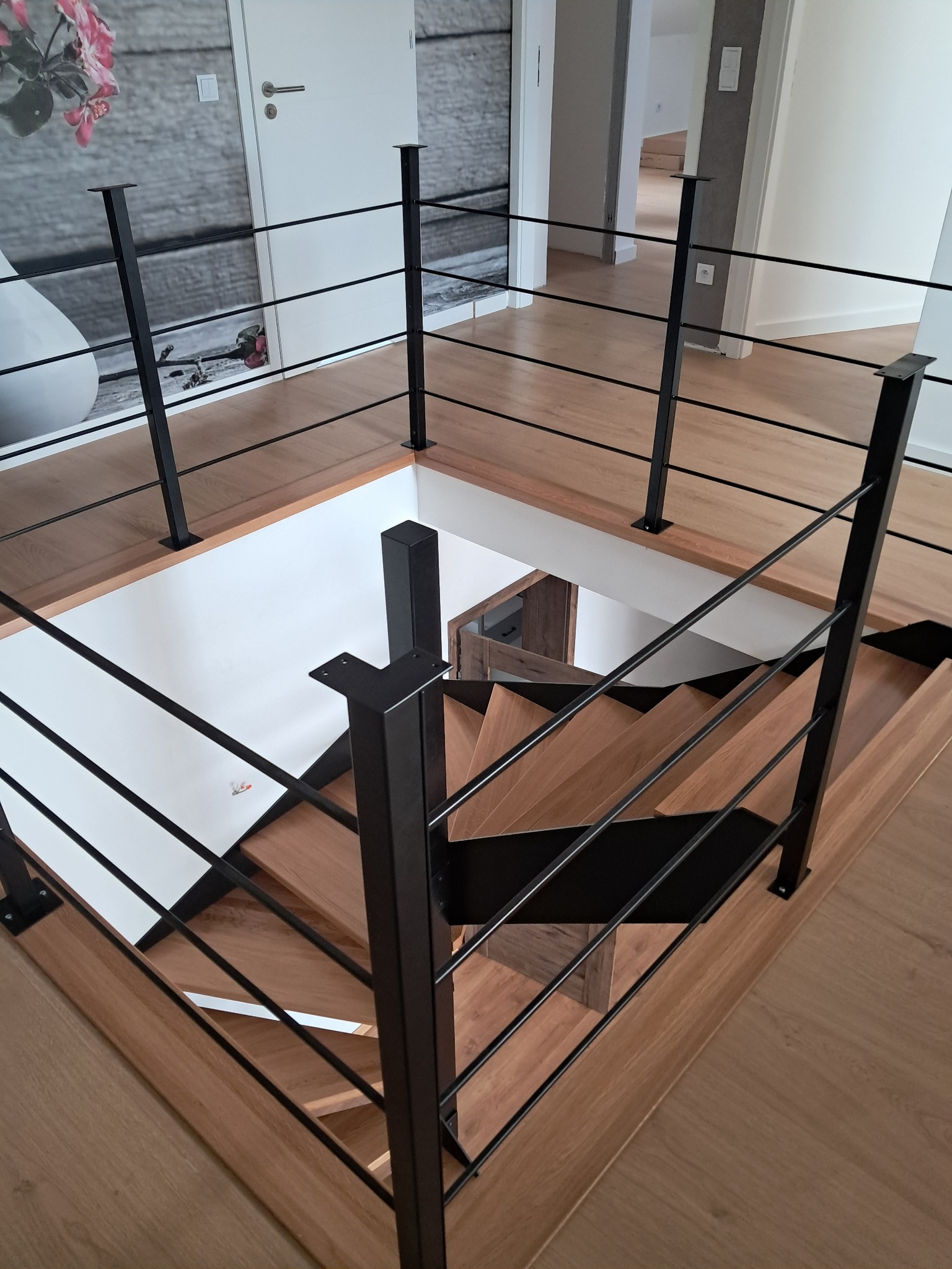 Wnętrze domu z drewnianymi schodami i czarną, minimalistyczną balustradą z poziomych prętów, widok z góry na klatkę schodową.