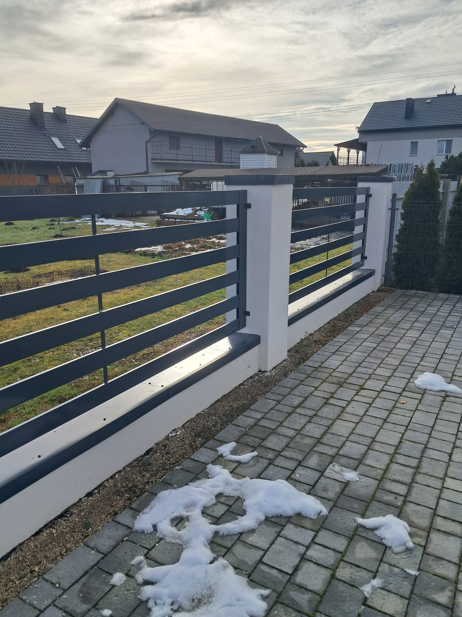 Nowoczesne ogrodzenie metalowe z poziomymi przęsłami w kolorze grafitowym, osadzone na białych, murowanych słupkach, z fragmentami topniejącego śniegu na kostce brukowej.