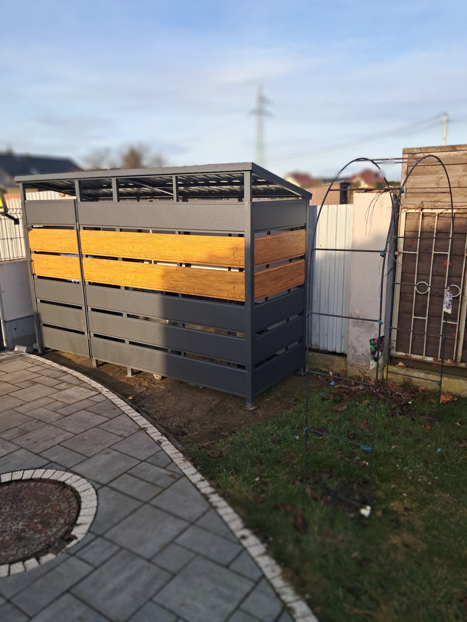 Szara obudowa śmietnikowa z drewnianymi panelami na posesji, widok z boku na tle trawnika i kostki brukowej.