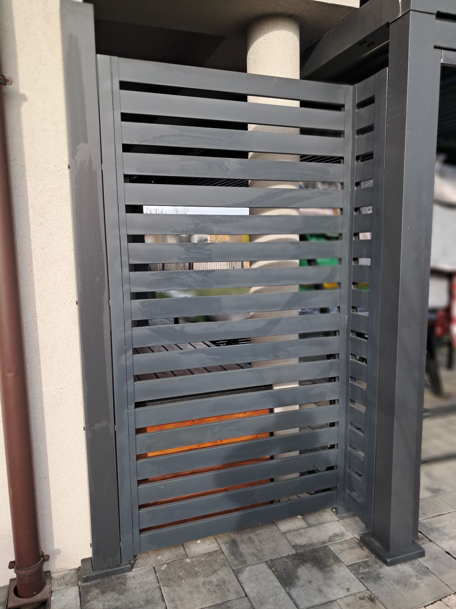 Nowoczesna, szara brama wjazdowa z poziomych profili, zamontowana na posesji z kostką brukową, widoczny fragment rynny w kolorze brązowym.