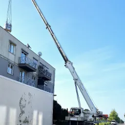 Dźwig podnosi pakiet materiałów na dach szarego bloku z balkonami, widoczny pracownik na dachu, obok zaparkowana ciężarówka z kolejnymi pakietami, błękitne niebo.