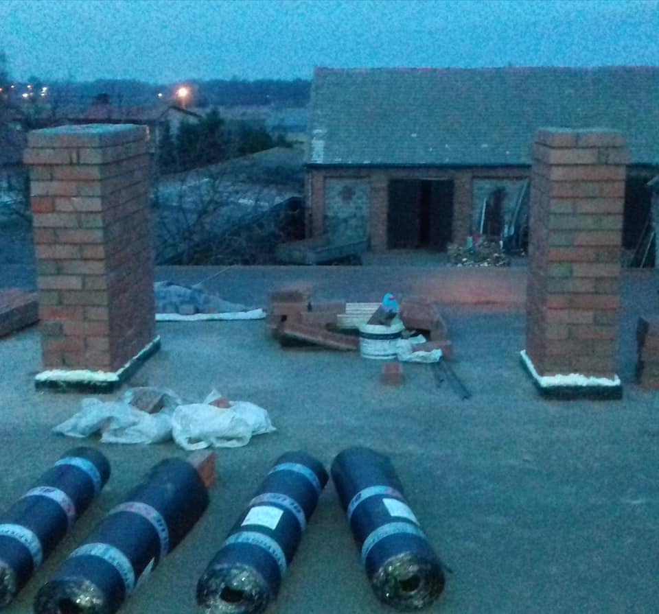 Widok z góry na płaski dach z czterema rolkami papy, dwoma kominami z cegły z uszczelnieniem u podstawy, oraz materiałami budowlanymi w tle, budynek gospodarczy.