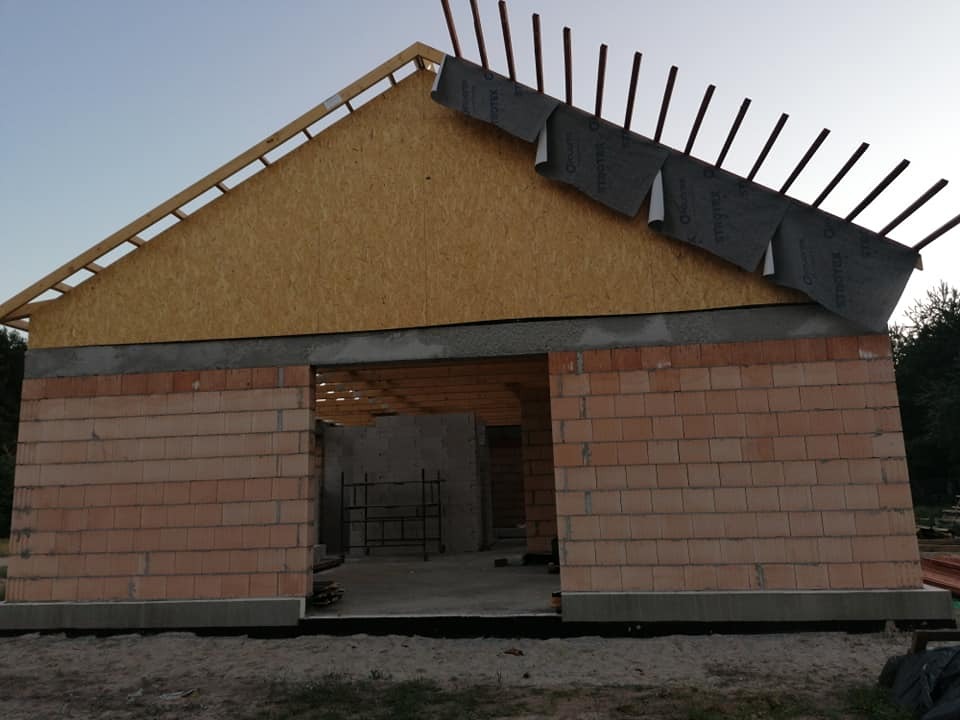 Budowa domu w trakcie, widoczne murowane ściany z czerwonej cegły, drewniana konstrukcja dachu z płytą OSB i częściowo ułożona membrana dachowa. Widoczne krokwie. Budynek w stanie surowym otwartym.