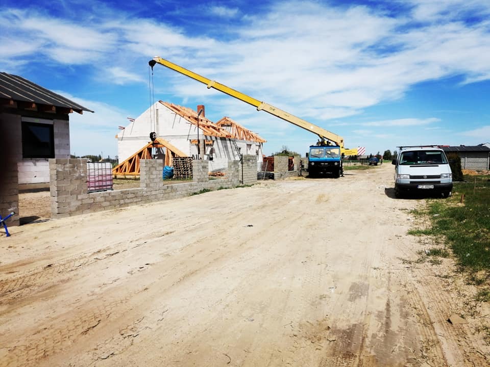 Montaż więźby dachowej z użyciem dźwigu samochodowego na placu budowy, widoczne elementy konstrukcyjne dachu i pustaki na ogrodzenie.