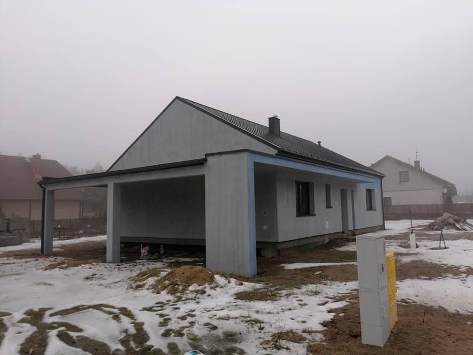 Nowoczesny dom jednorodzinny z płaskim, ciemnym dachem i otwartą wiatą garażową, widoczny w pochmurny dzień z resztkami śniegu na ziemi.