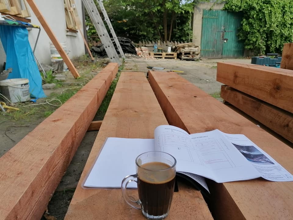 Przygotowanie do prac dekarskich: Plany na otwartej książce i kawa na belkach drewnianych w plenerze, w tle drabina, palety i zielone drzwi do budynku.