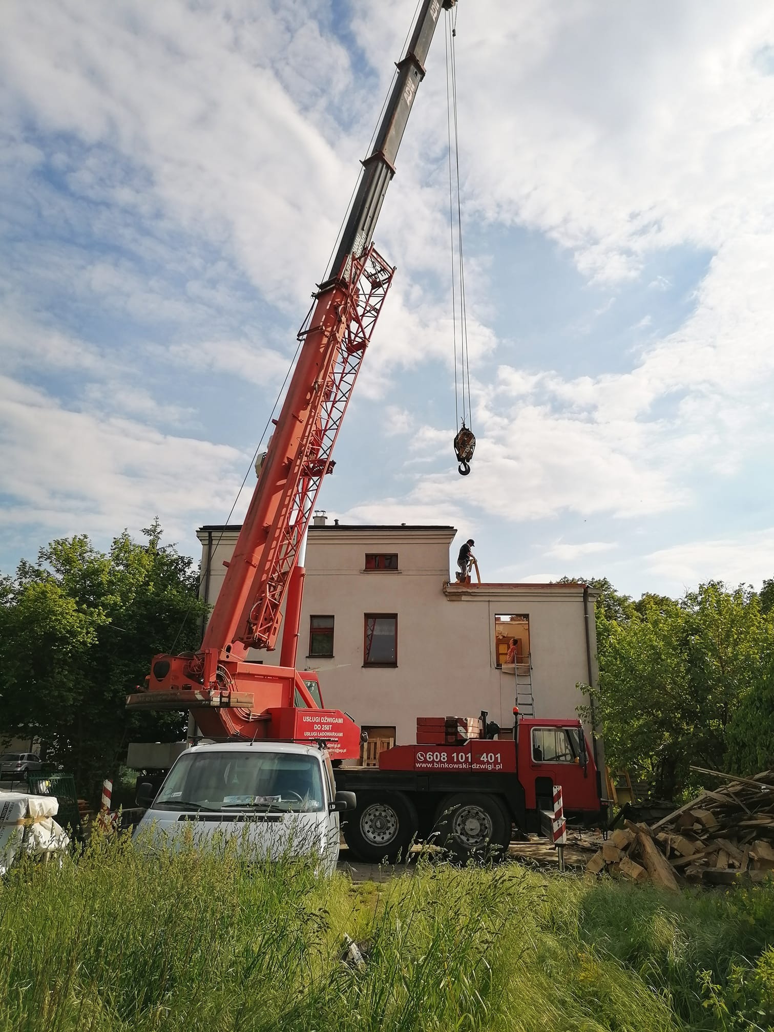 Prace na dachu dwupiętrowego budynku z użyciem dźwigu, widoczny pracownik na dachu i osoba w oknie, gruz i resztki materiałów budowlanych na trawie obok budynku.