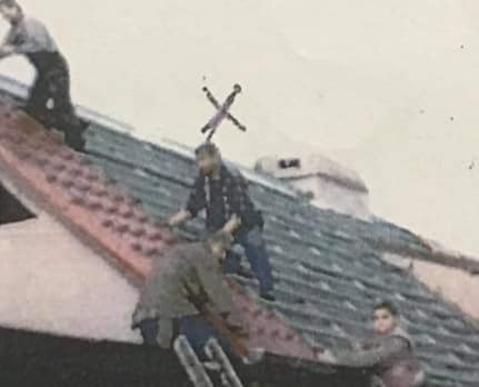 Czterech mężczyzn pracuje na dachu z czerwonej i zielonej dachówki, jeden z nich na drabinie, widoczny komin w tle.