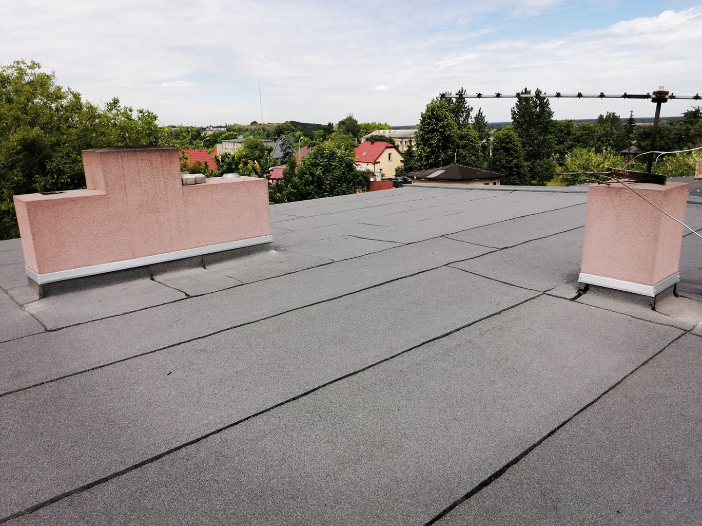 Płaski dach pokryty papą termozgrzewalną z dwoma kominami w kolorze różowym, widok na okoliczne budynki i zieleń w tle.