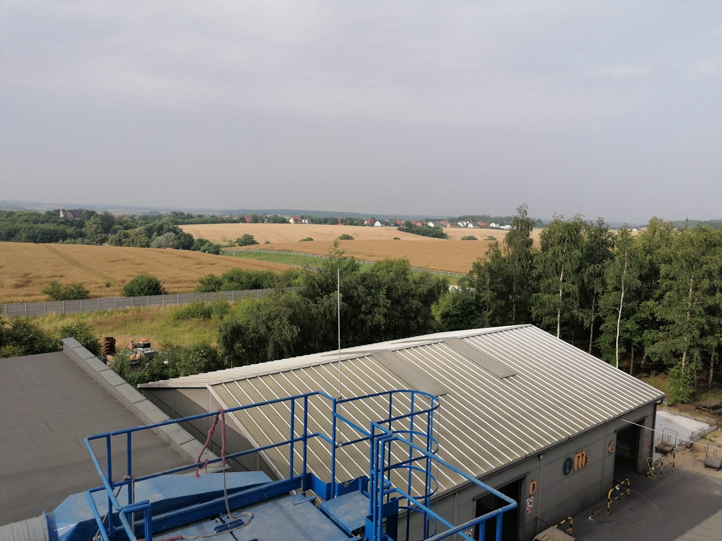 Widok z wysokości na budynek z dachem trapezowym, otoczony polami i lasami w oddali, z elementami niebieskiej konstrukcji na pierwszym planie.