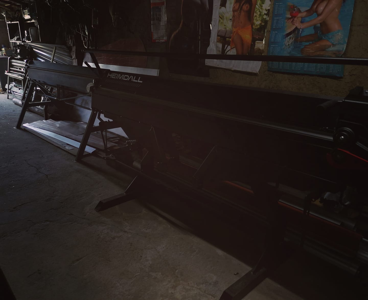 Długa, czarna giętarka do blachy marki Heimdall w ciemnym warsztacie, z widocznymi arkuszami blachy i kalendarzem na ścianie w tle.