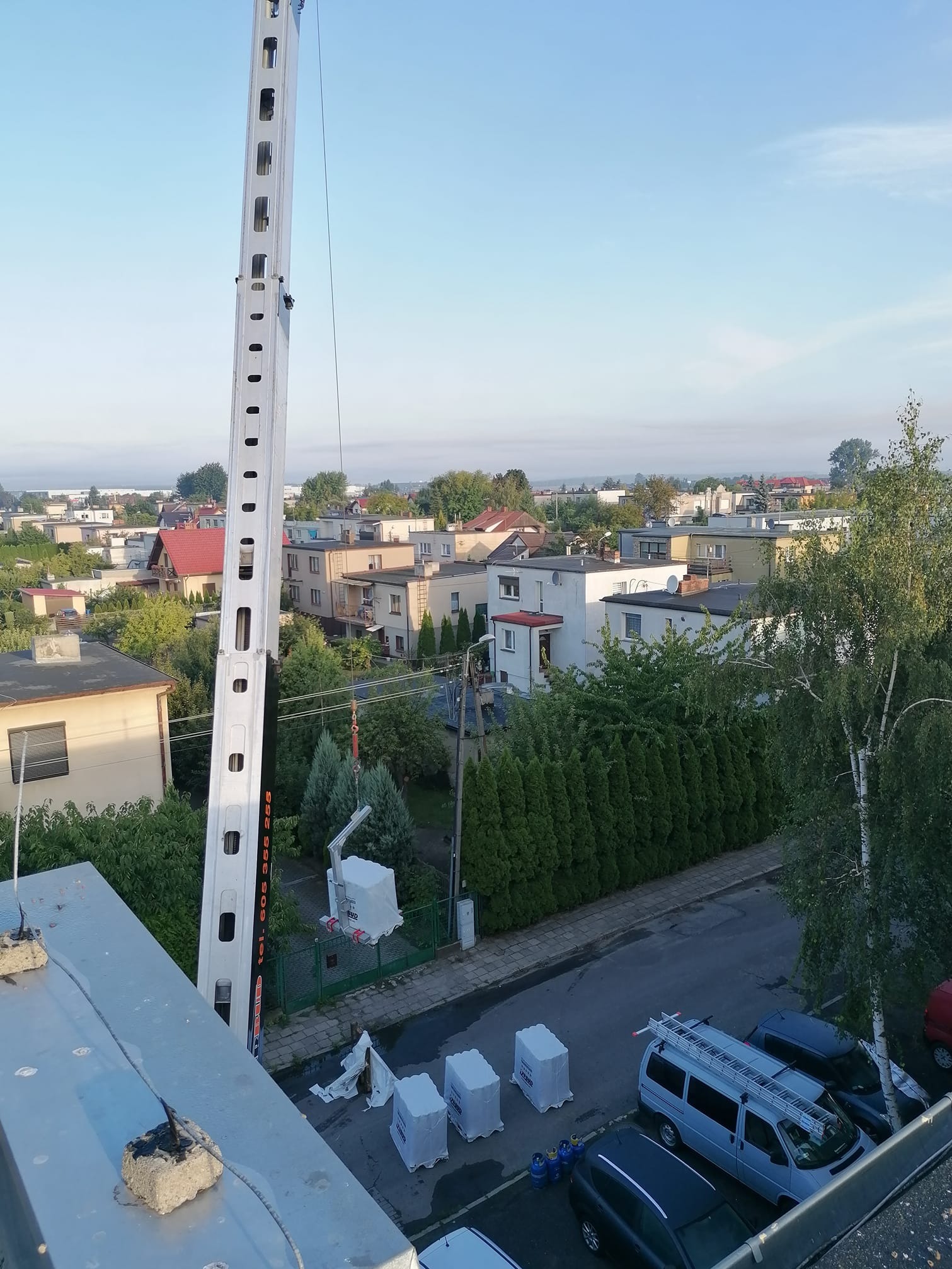 Widok z góry na podnośnik koszowy z zamontowanym pakunkiem, stojący na tle zabudowy miejskiej i zaparkowanych samochodów, sugerujący transport materiałów na dach.