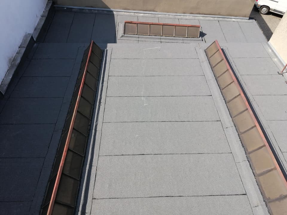 Płaski dach pokryty papą termozgrzewalną z świetlikami dachowymi w brązowych obramowaniach. Widoczny fragment budynku w tle oraz samochód dostawczy.