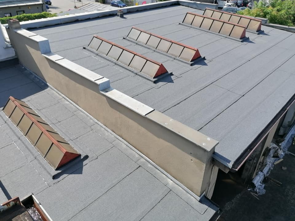 Widok z góry na płaski dach pokryty papą termozgrzewalną z czterema świetlikami dachowymi i attyką z elementami blacharskimi.