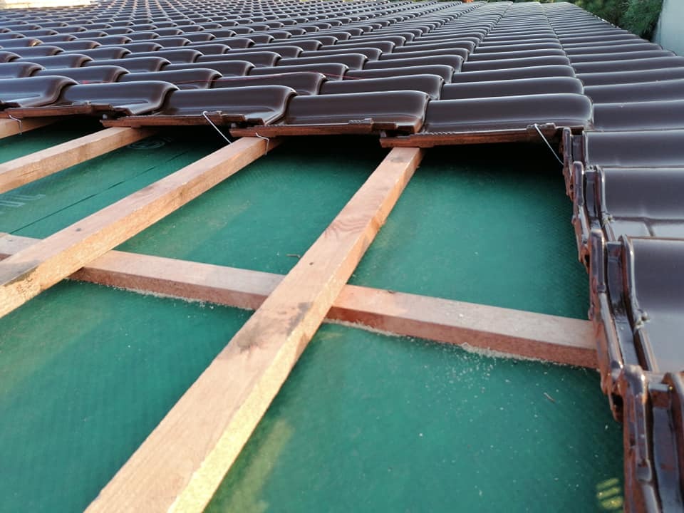 Montaż brązowej dachówki ceramicznej na konstrukcji drewnianej z zieloną membraną dachową, widok z góry na częściowo pokryty dach.