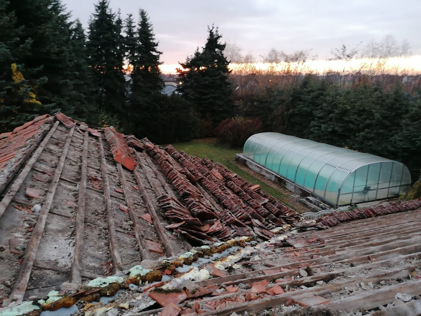 Demontaż starej dachówki ceramicznej, widoczne drewniane elementy konstrukcji dachu, resztki zaprawy i pianki montażowej, w tle zielony ogród z zadaszonym basenem i drzewami o zachodzie słońca.