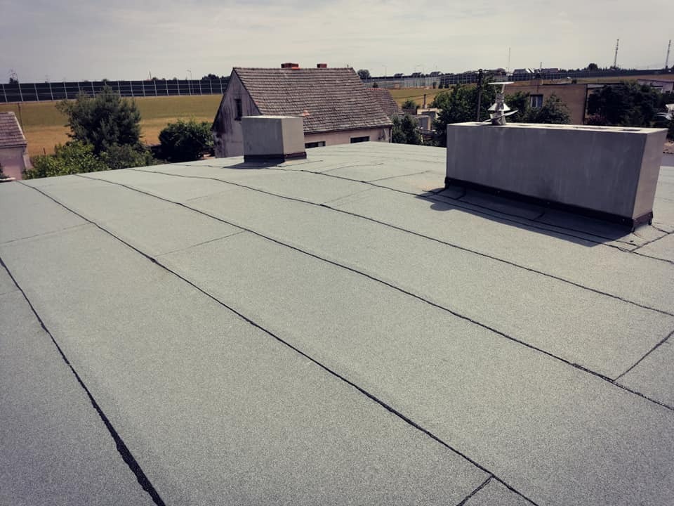 Szczelny, płaski dach pokryty papą termozgrzewalną z widocznymi kominami wentylacyjnymi i w tle zabudowa jednorodzinna.