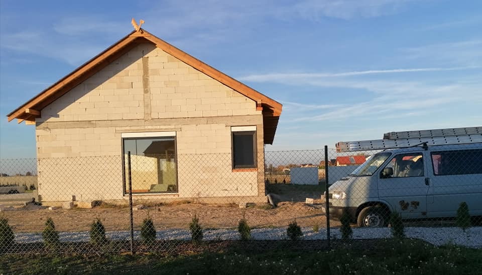 Surowy stan domu w trakcie budowy z widoczną drewnianą konstrukcją dachu i zaparkowanym obok busem z drabinami na dachu.