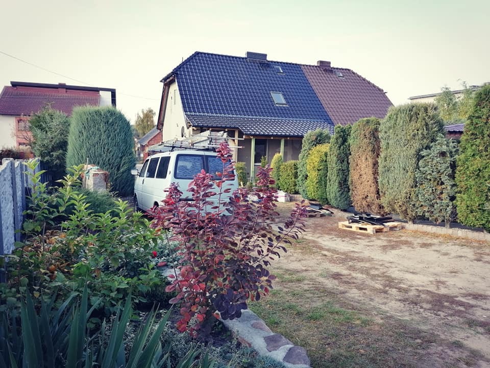 Dom z dachem pokrytym w połowie ciemną dachówką, w połowie brązową, widoczny samochód dostawczy z drabiną na dachu oraz palety z materiałami budowlanymi w ogrodzie.