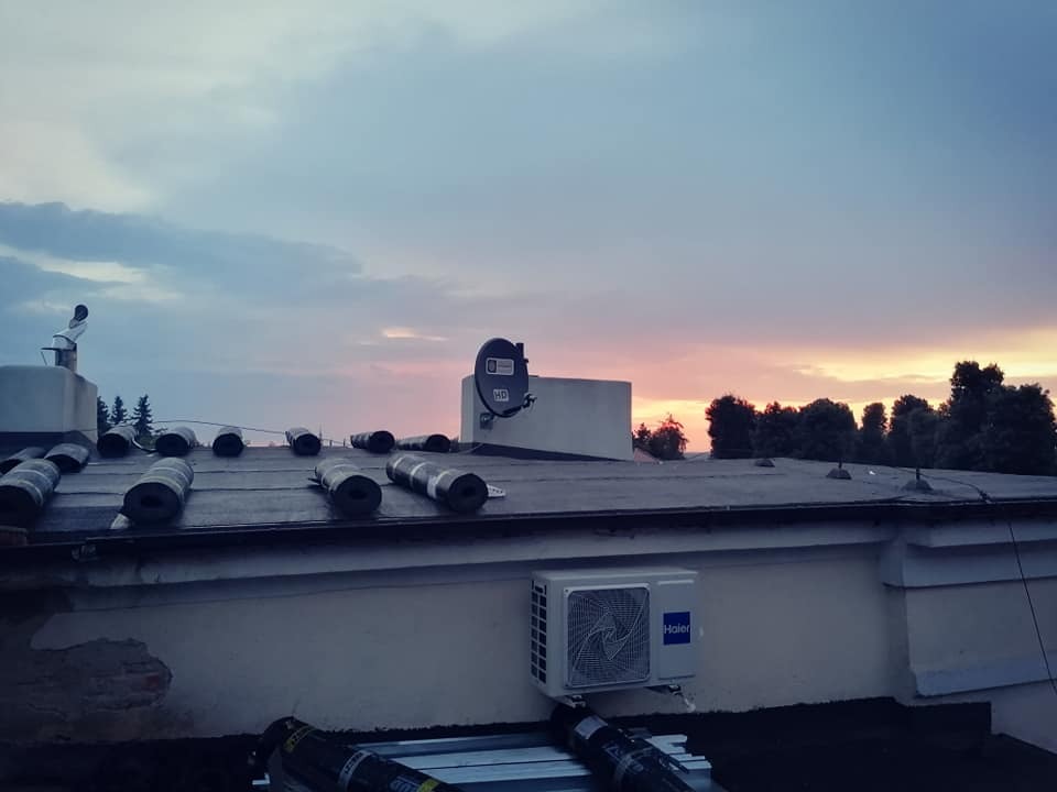 Przygotowany do położenia dach pokryty papą na tle zachodzącego słońca, widoczna antena satelitarna i klimatyzator na elewacji.