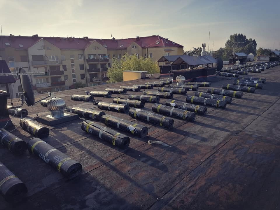 Przygotowanie do hydroizolacji dachu płaskiego: rozłożone rolki papy bitumicznej IZOBUD na czarnej powierzchni, w tle budynki mieszkalne i wirnik wentylacyjny.