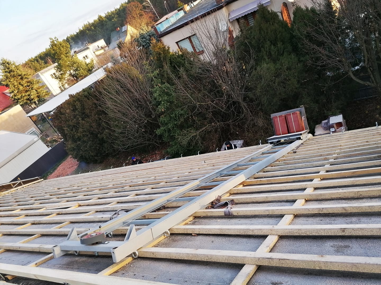 Konstrukcja dachu z widocznym podnośnikiem do dachówki, przygotowana do krycia, w tle zabudowa jednorodzinna i zieleń.