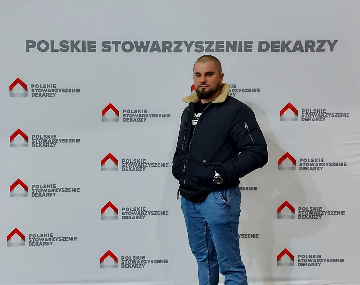 Mężczyzna pozuje na tle ściany z logo Polskiego Stowarzyszenia Dekarzy w Śremie.