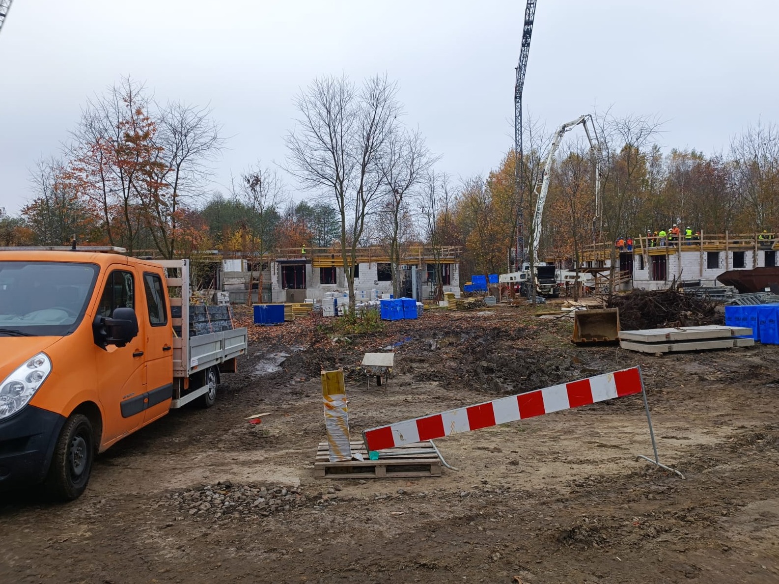 Budowa osiedla domów jednorodzinnych w stanie surowym, widoczny pomarańczowy samochód dostawczy z ładunkiem, ekipa budowlana na rusztowaniach, betonowa pompa i tymczasowe zabezpieczenie budowy...