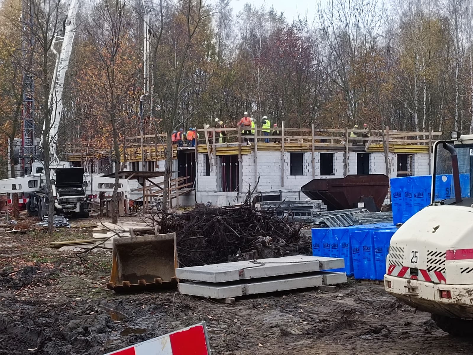 Budowa parterowego budynku z bloczków betonowych w trakcie realizacji, widoczni robotnicy w kaskach i kamizelkach na rusztowaniu, dźwig do betonu, wykopany teren, elementy szalunków i materiały...