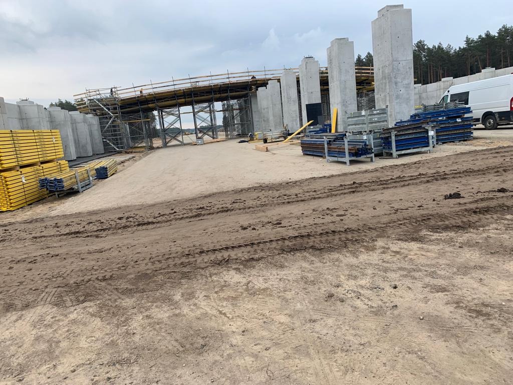 Budowa wiaduktu: surowe betonowe filary i szalunki, widoczne rusztowania i materiały budowlane na placu budowy, w tle las.