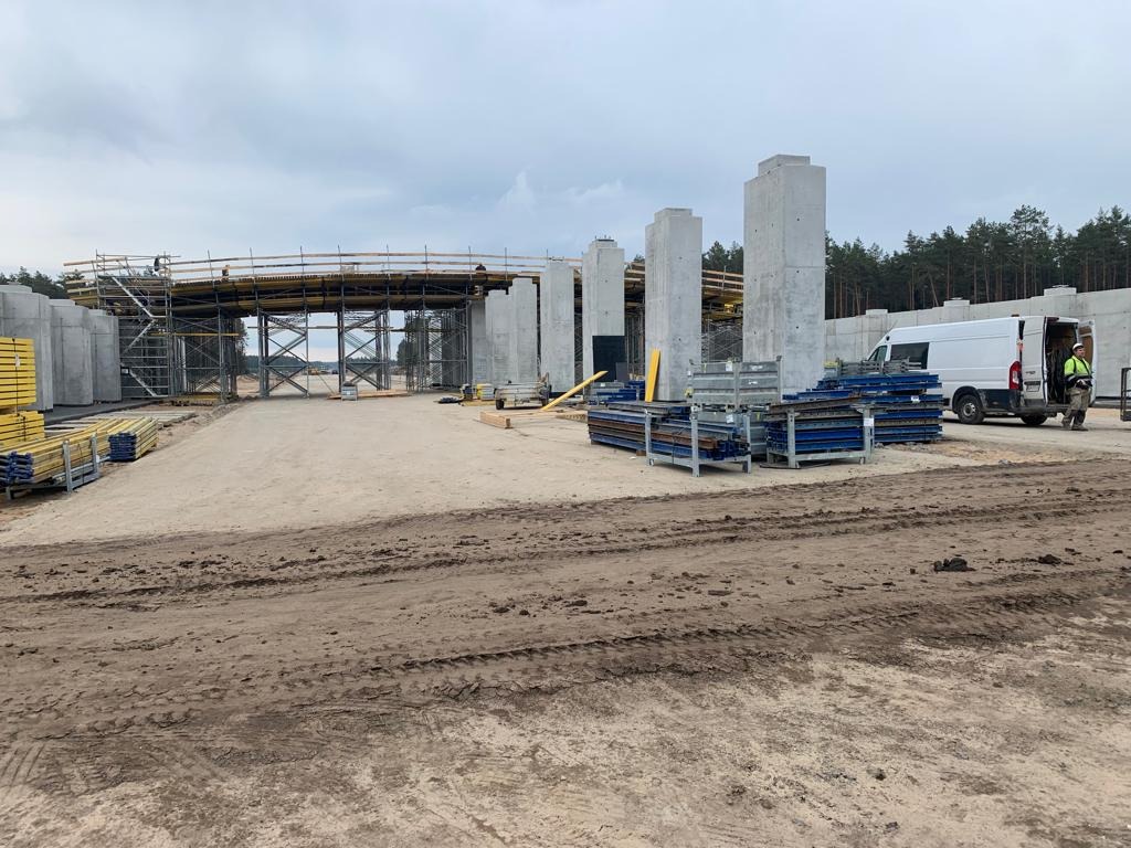 Budowa wiaduktu: betonowe filary, stalowe rusztowania, transportowy van i pracownik w odblaskowej kamizelce na placu budowy.