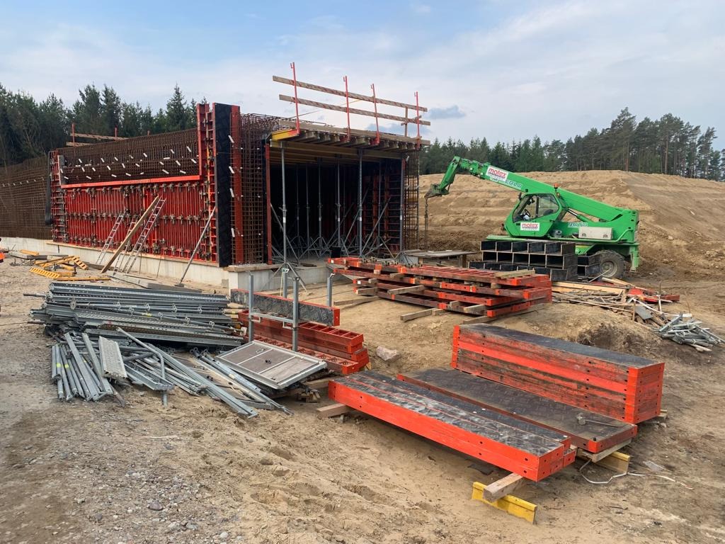 Konstrukcja żelbetowa w trakcie budowy: szalunek zbrojony stalą, zielony podnośnik teleskopowy i stosy drewnianych belek na placu budowy w słoneczny dzień.