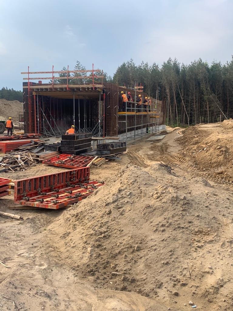 Budowa żelbetowej konstrukcji przepustu drogowego z użyciem szalunków i zbrojenia, widoczni pracownicy w kaskach i kamizelkach odblaskowych, teren budowy z hałdami piasku.