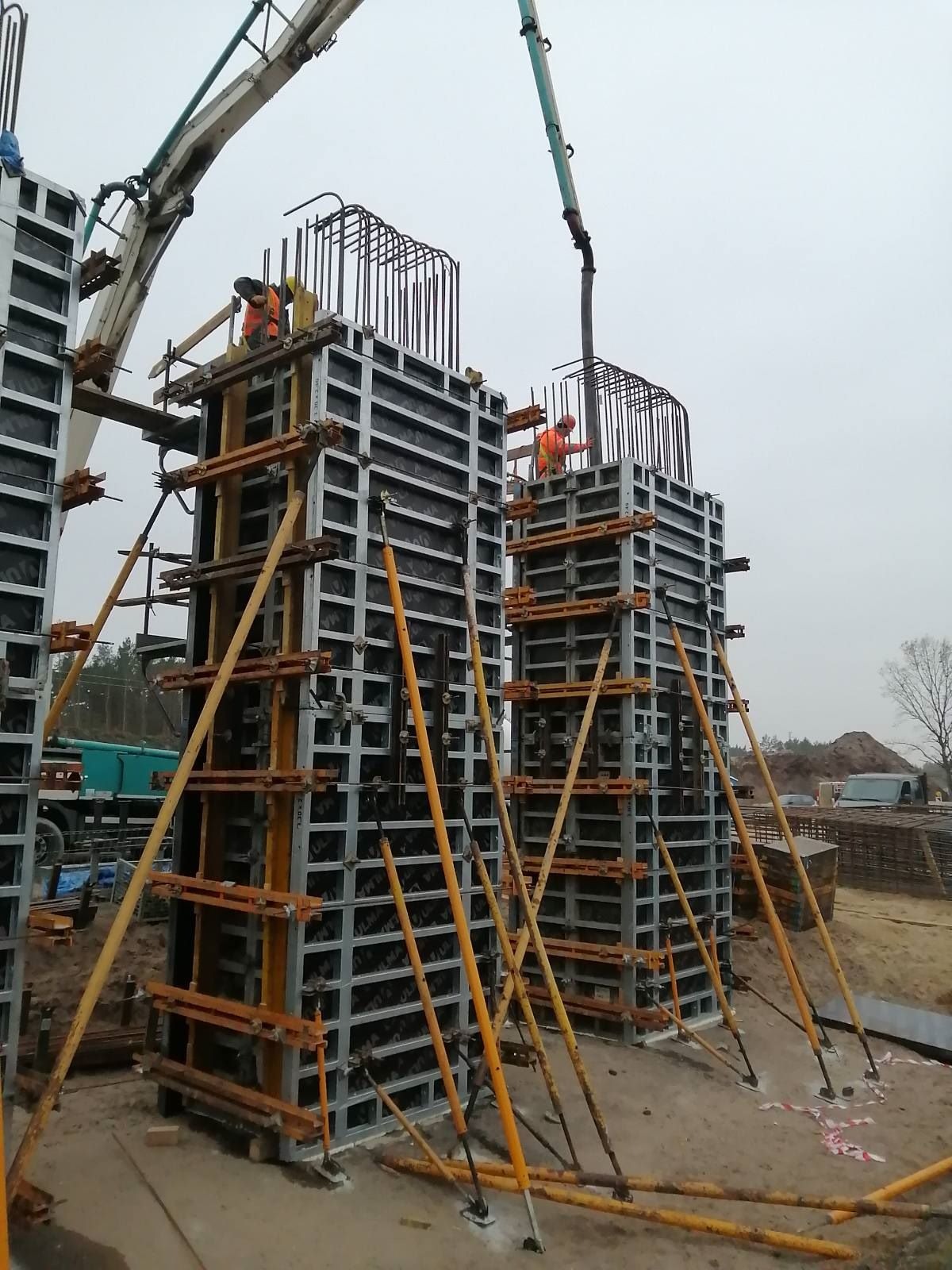 Wylewanie betonu do szalunków kolumn zbrojeniowych na placu budowy, widoczni pracownicy w pomarańczowych kamizelkach i betonowóz z pompą.