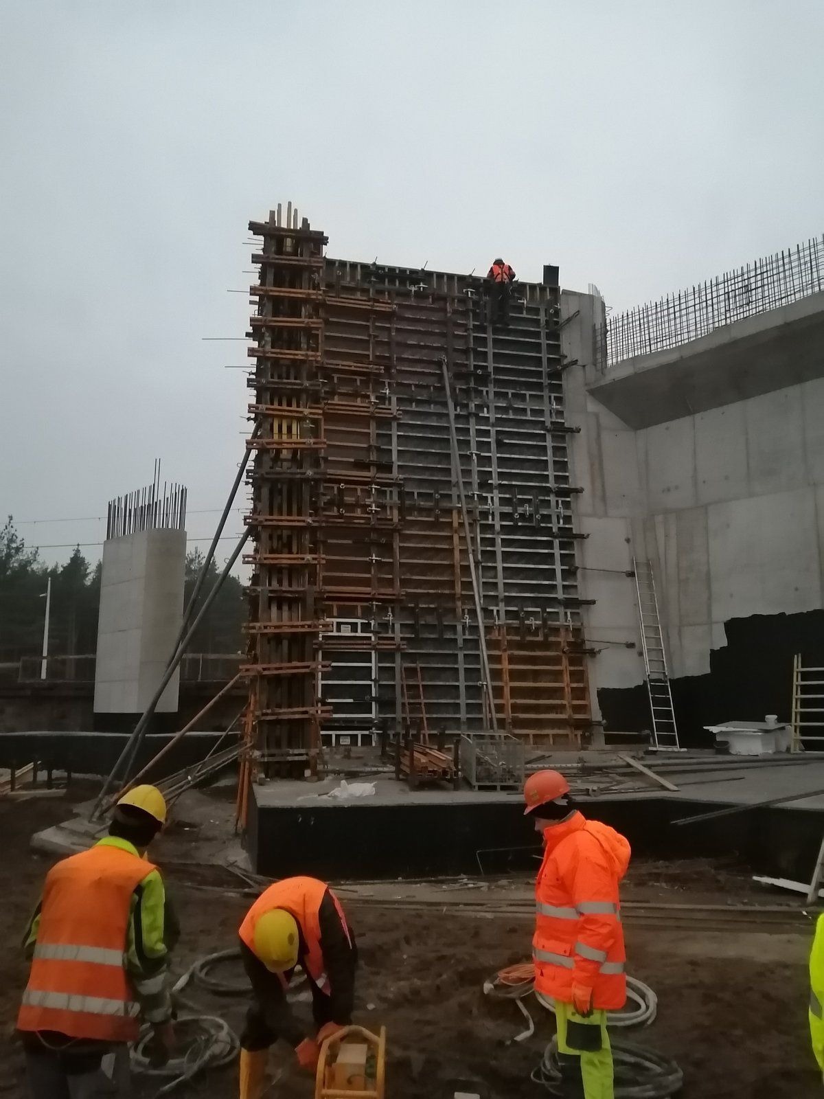 Wysoka ściana szalunkowa z drewna i metalu na placu budowy, widoczni robotnicy w odblaskowych ubraniach i kaskach, na górze ściany jeden robotnik, w tle betonowy element konstrukcyjny i zalesiony...