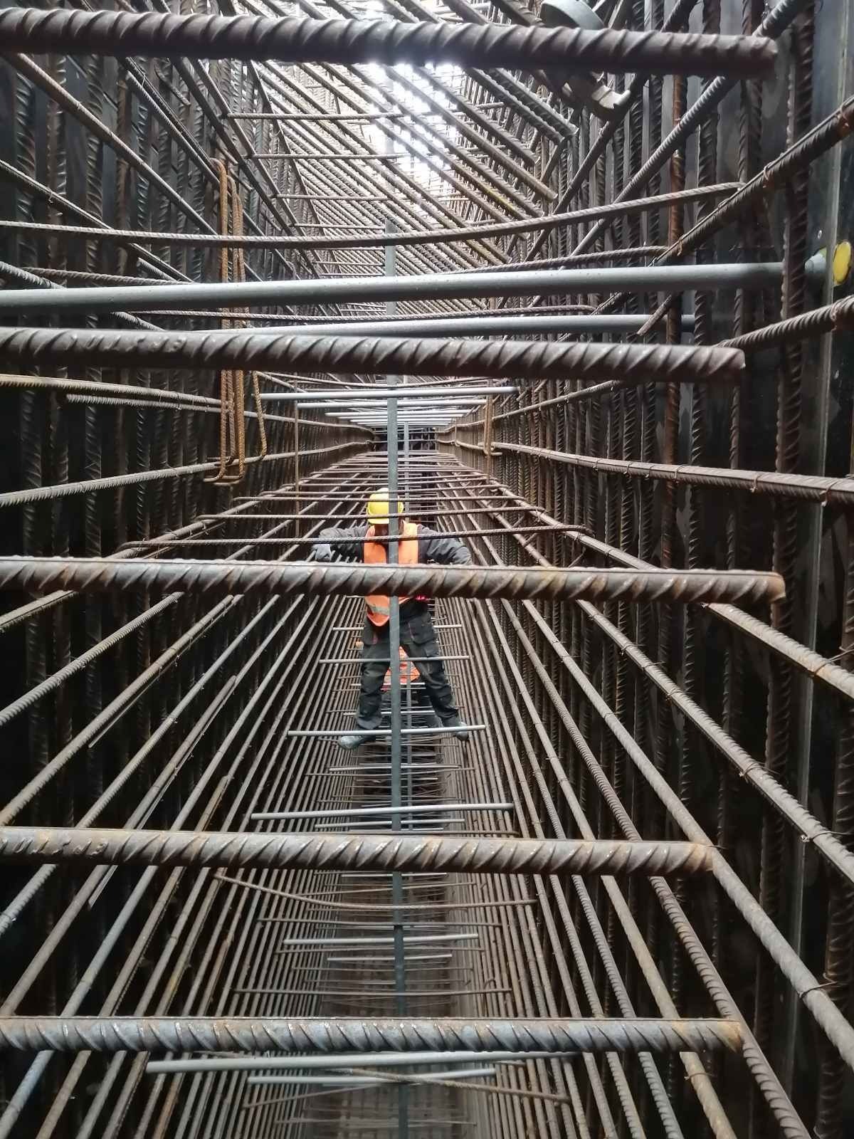 Pracownik w pomarańczowej kamizelce i żółtym kasku stoi na metalowej konstrukcji z prętów zbrojeniowych wewnątrz szalunku, widok z dołu.
