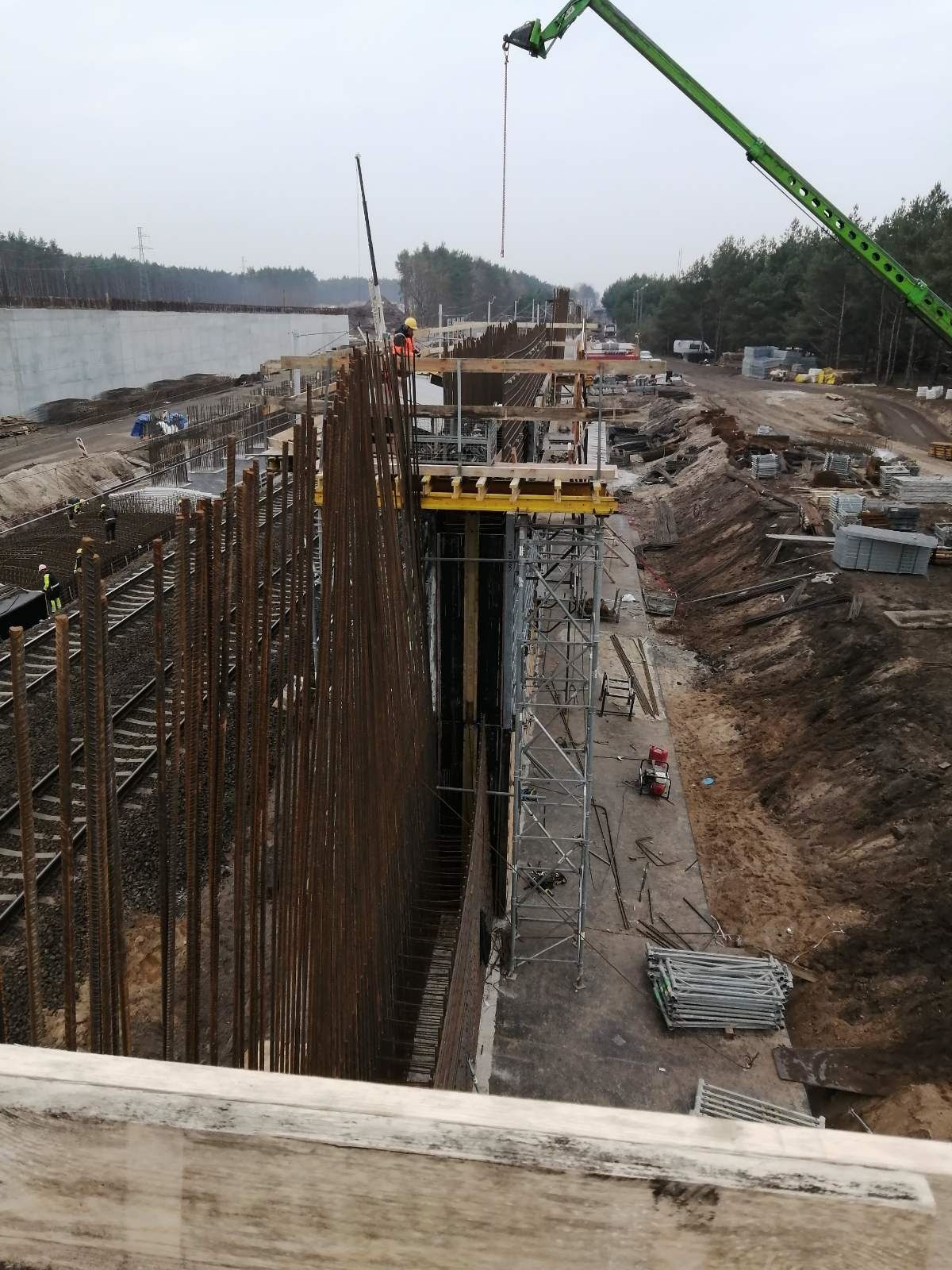 Budowa wiaduktu: widok na pionowe pręty zbrojeniowe, szalunki, rusztowania oraz dźwig na placu budowy w pochmurny dzień.
